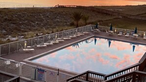 Zona de piscina con sillones de descanso y una terraza de madera con vistas a una playa al atardecer.