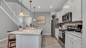 A modern kitchen with white cabinetry, a central island, stainless steel appliances, pendant lighting, and a staircase leading to another level. - Luxury Home 7 Bedrooms Near Disney! 7 Villa by RedAwning (Kissimmee)