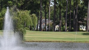 Golfkenttä, jossa on suihkulähde, harjoittelualue lyhyitä lyöntejä varten ja korkeita puita taustalla.