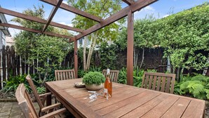 A wooden table with chairs, a potted plant, and a bottle of wine in an outdoor garden setting. - Valley Palms - Mt Maunganui Holiday Home (Mt Maunganui)