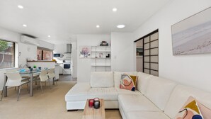 A modern living room with a white sofa, a glass dining table, and a kitchen in the background. - Valley Palms - Mt Maunganui Holiday Home (Mt Maunganui)