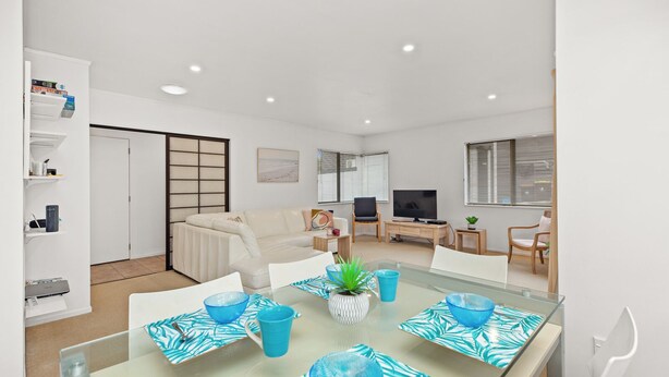 A modern living room with a glass dining table, white sofas, a TV, and a bookshelf. - Valley Palms - Mt Maunganui Holiday Home (Mt Maunganui)