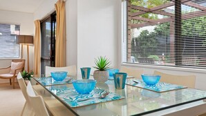A modern dining area with a glass table, blue glassware, and a potted plant. - Valley Palms - Mt Maunganui Holiday Home (Mt Maunganui)