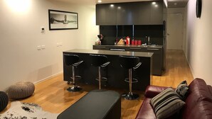 Une cuisine moderne dotée d’un îlot central et de tabourets de bar, un coin repas avec un canapé en cuir et un pouf, et un design minimaliste agrémenté d’un éclairage encastré.