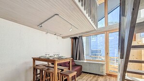 A dining area with a wooden table and chairs, a window with curtains, and a view of the outside. - Les Genepis (Val Claret) by Interhome (Tignes)
