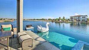 An infinity pool with a swan float, outdoor seating, and a view of a large house across the water.