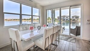 A modern dining area with a white table and chairs, large windows, and a view of a swimming pool and houses.