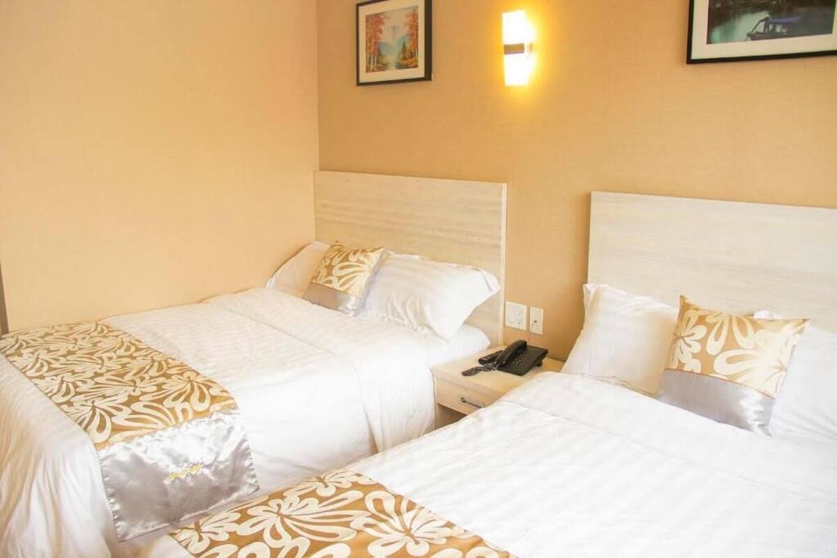 Hotellihuone, jossa on kaksi sänkyä. Kummassakin sängyssä on valkoiset vuodevaatteet ja kuviollinen peitto. Huoneessa on yöpöytä, jossa on puhelin, sekä seinään kiinnitetty valaisin.