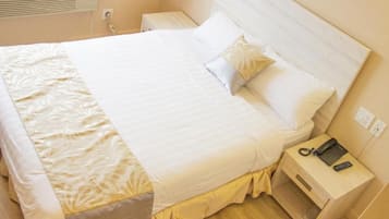 Una cama bien hecha con sábanas blancas y una almohada decorativa, una mesita de noche con teléfono y control remoto, y un equipo de aire acondicionado instalado en la pared.