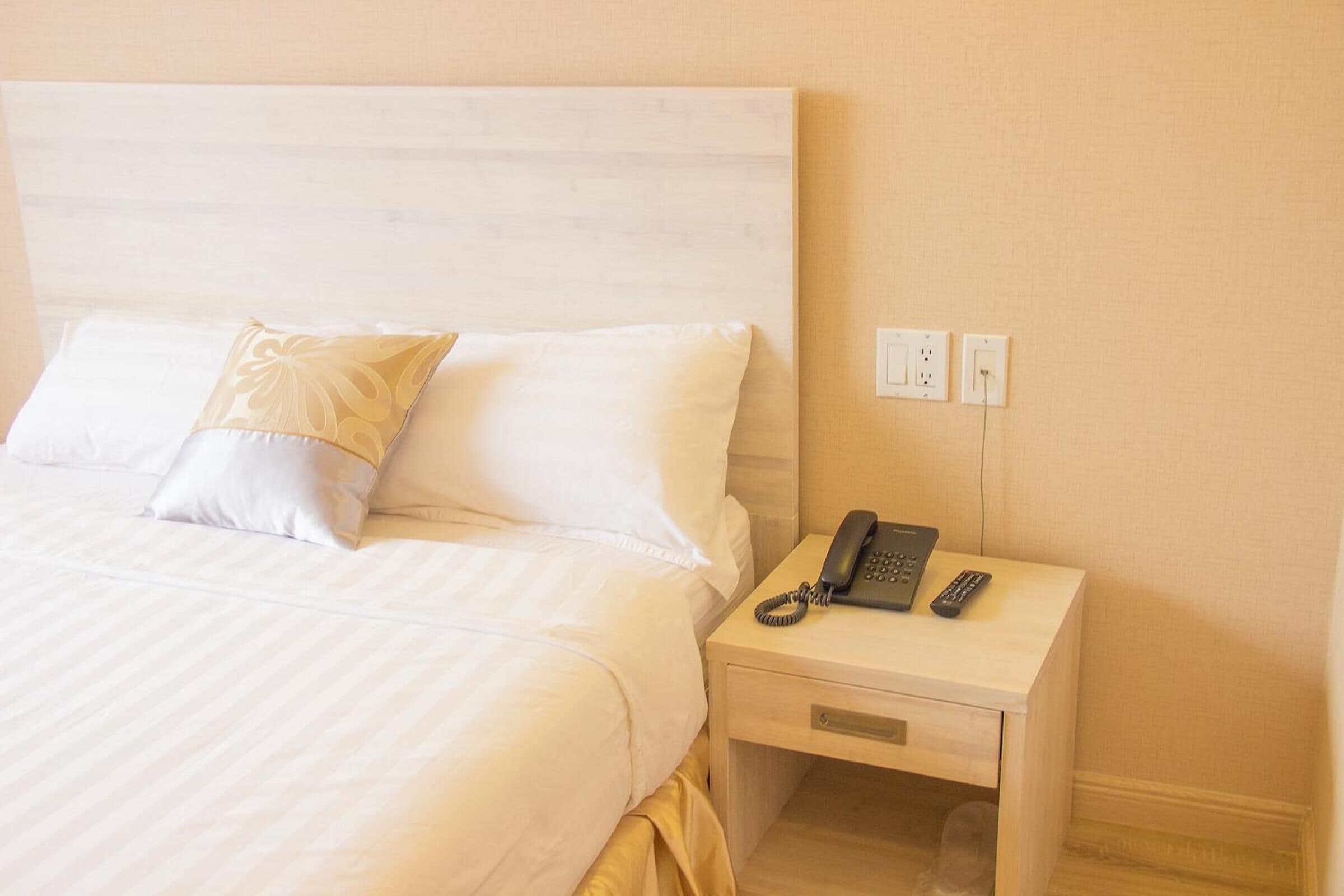 A neatly made bed with white linens and a single decorative pillow, a bedside table with a telephone and remote controls, and a wall socket.