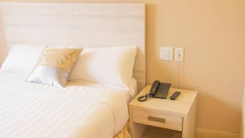 A neatly made bed with white linens and a single decorative pillow, a bedside table with a telephone and remote controls, and a wall socket.