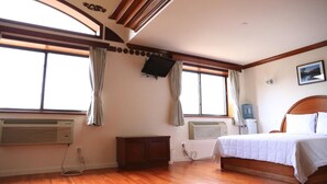 Quarto com piso de madeira, cama com roupa de cama branca, duas janelas com cortinas e ar condicionado instalado.