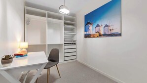 Ett rum med ett vitt skrivbord, en stol och en bokhylla. På väggen hänger en tavla med motiv av väderkvarnar, och på skrivbordet står en lampa.