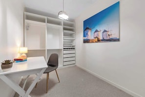Ein Zimmer mit weißem Schreibtisch, Stuhl und Bücherregal. An der Wand hängt ein Windmühlenbild und auf dem Schreibtisch steht eine Lampe.