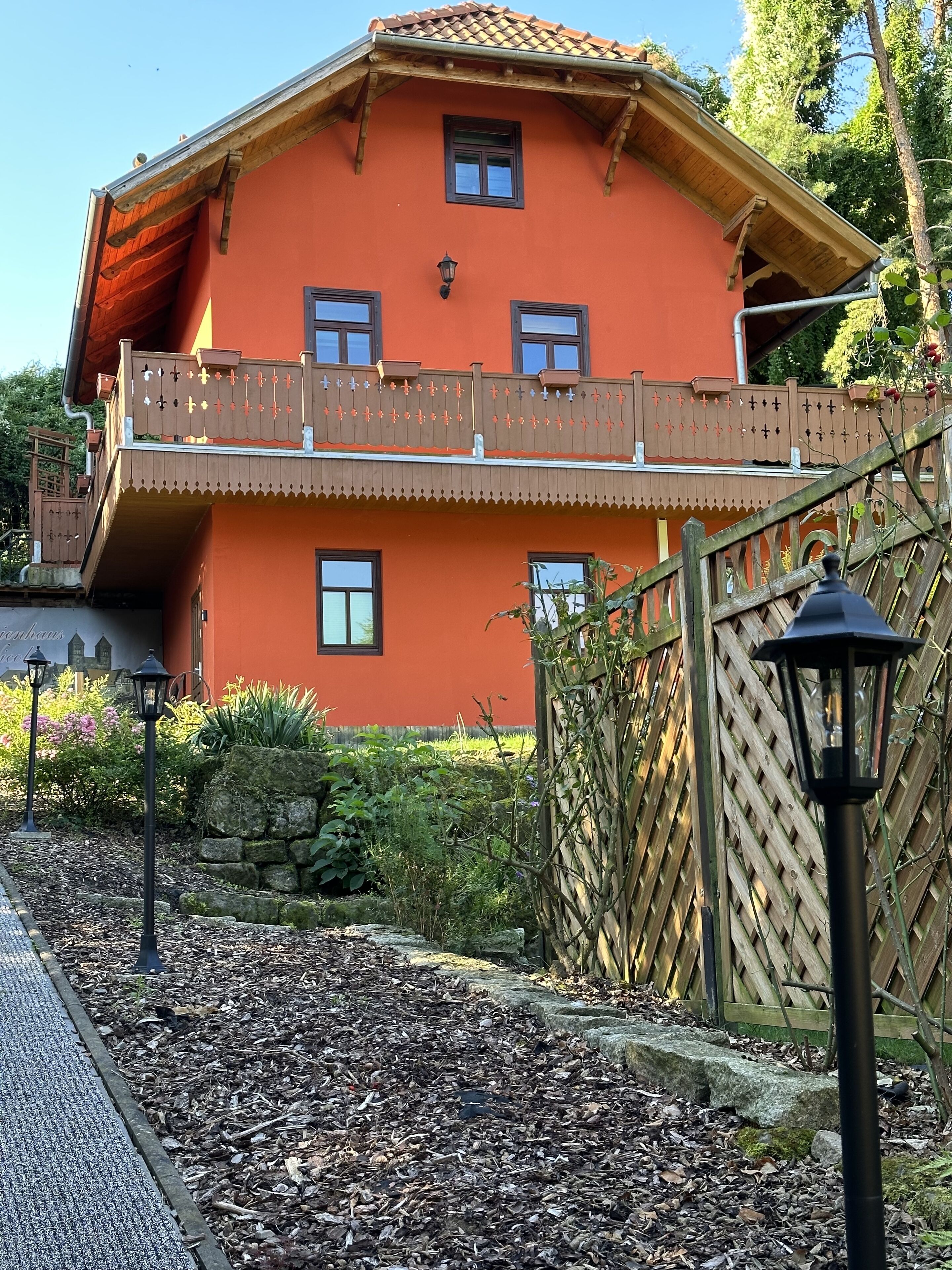 Une maison à deux étages dotée d’un balcon, entourée d’un jardin avec des murs en pierre et des plantes.