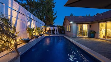 Relax by the serene pool surrounded by lush greenery and a charming house with a covered patio.