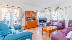 Ein Wohnzimmer mit einem blauen Sofa, einem violetten Sofa, einem Holzschrank, einem Fernseher und einem Fenster mit Vorhängen.