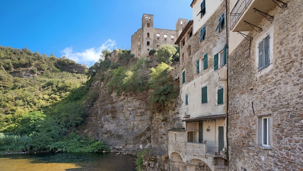 Un bâtiment en pierre doté de balcons donnant sur une rivière, un château perché sur une colline et une végétation luxuriante.