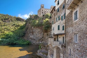 Ein Steinhaus mit Balkonen, die auf einen Fluss blicken, eine Burg auf einem Hügel und üppiges Grün.