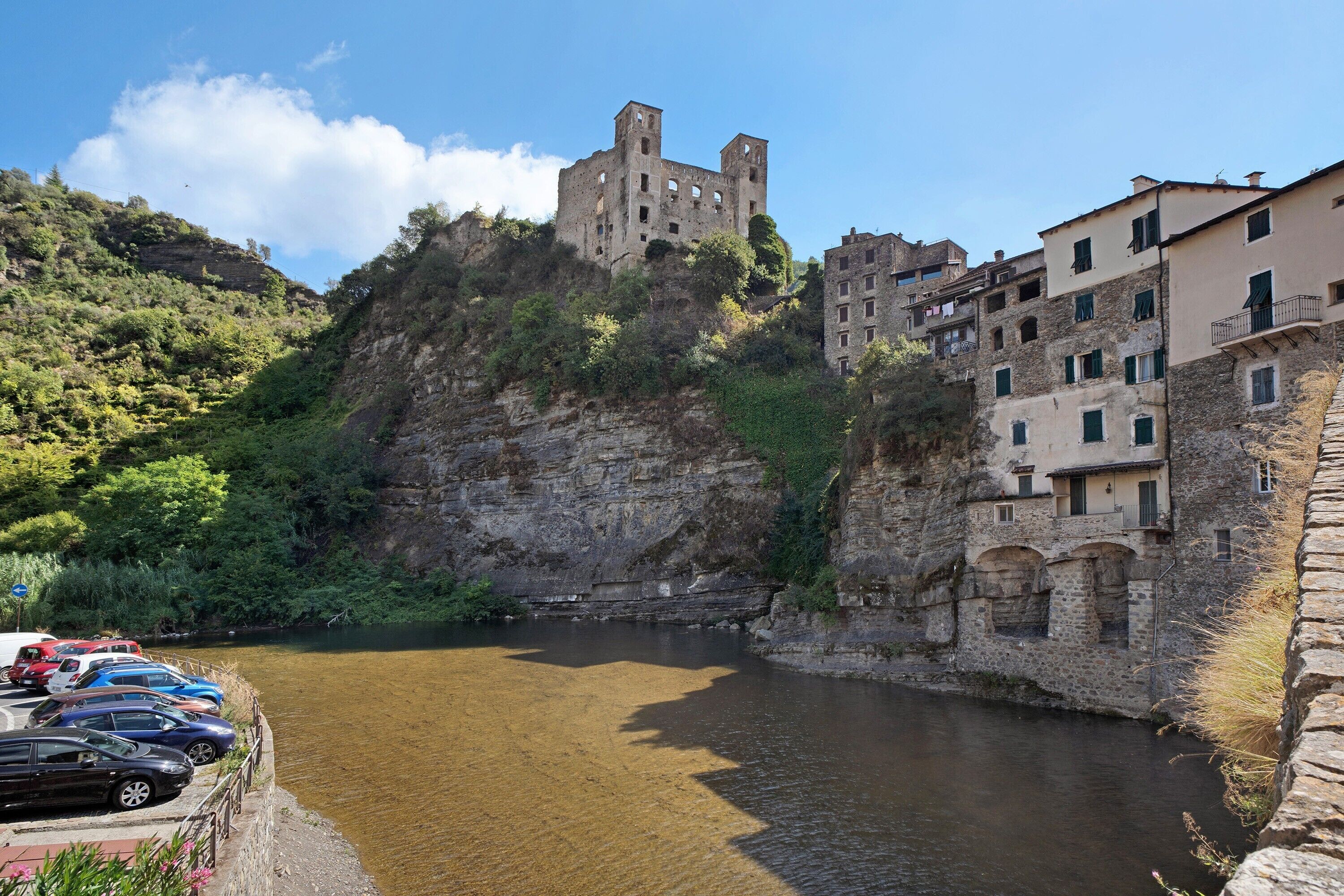 Ein historisches Steinhaus mit einem Fluss davor, parkenden Autos und einem wolkenlosen Himmel.