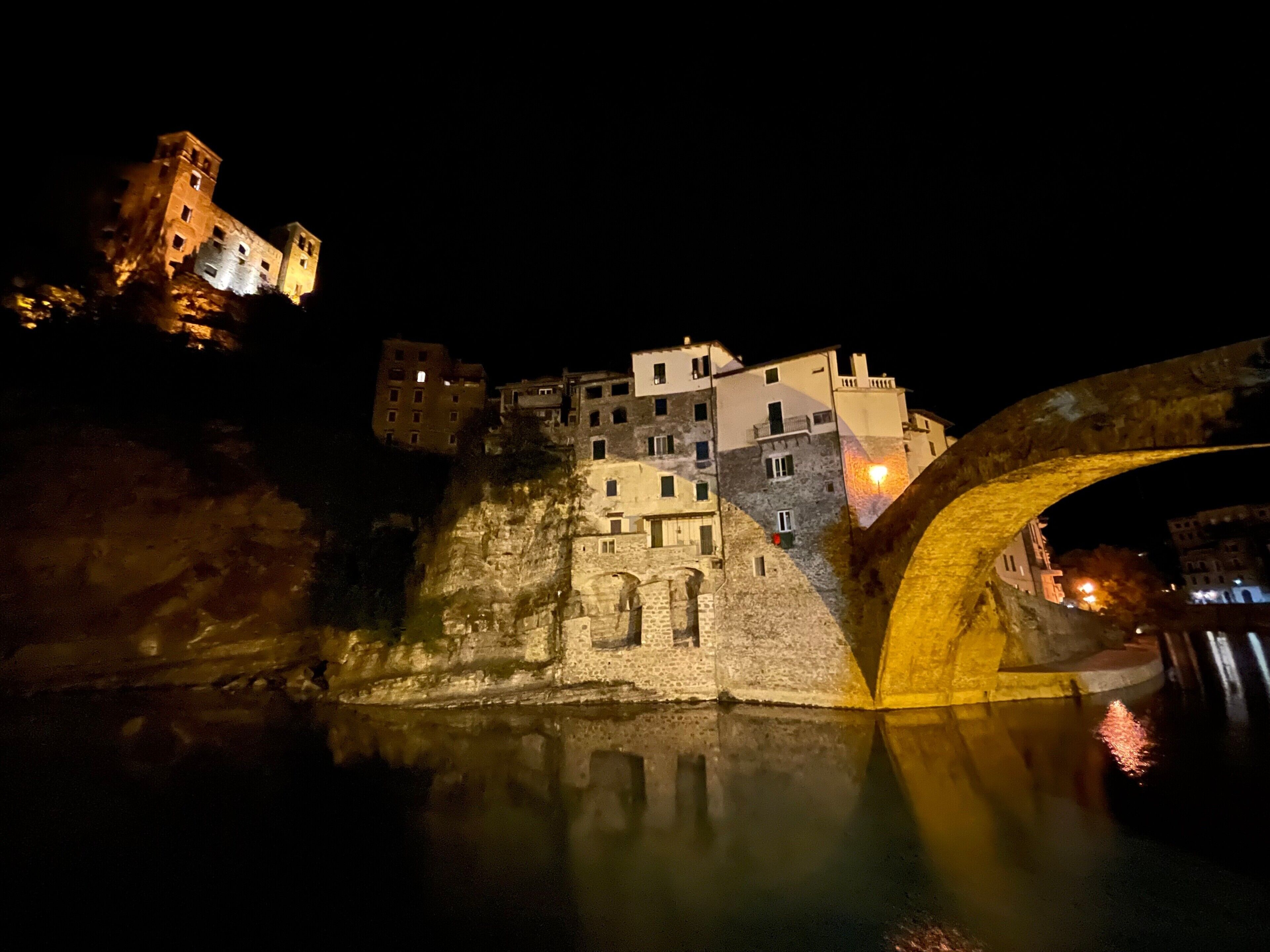 A historic stone bridge with a reflection in the water, leading to a town with illuminated buildings.