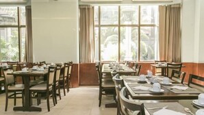 Uma sala de jantar com mesas e cadeiras, janelas amplas com cortinas e aparelhos de ar condicionado.