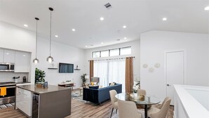 A modern kitchen with a central island, a dining area with a glass table, and a living space with a sofa and curtains.