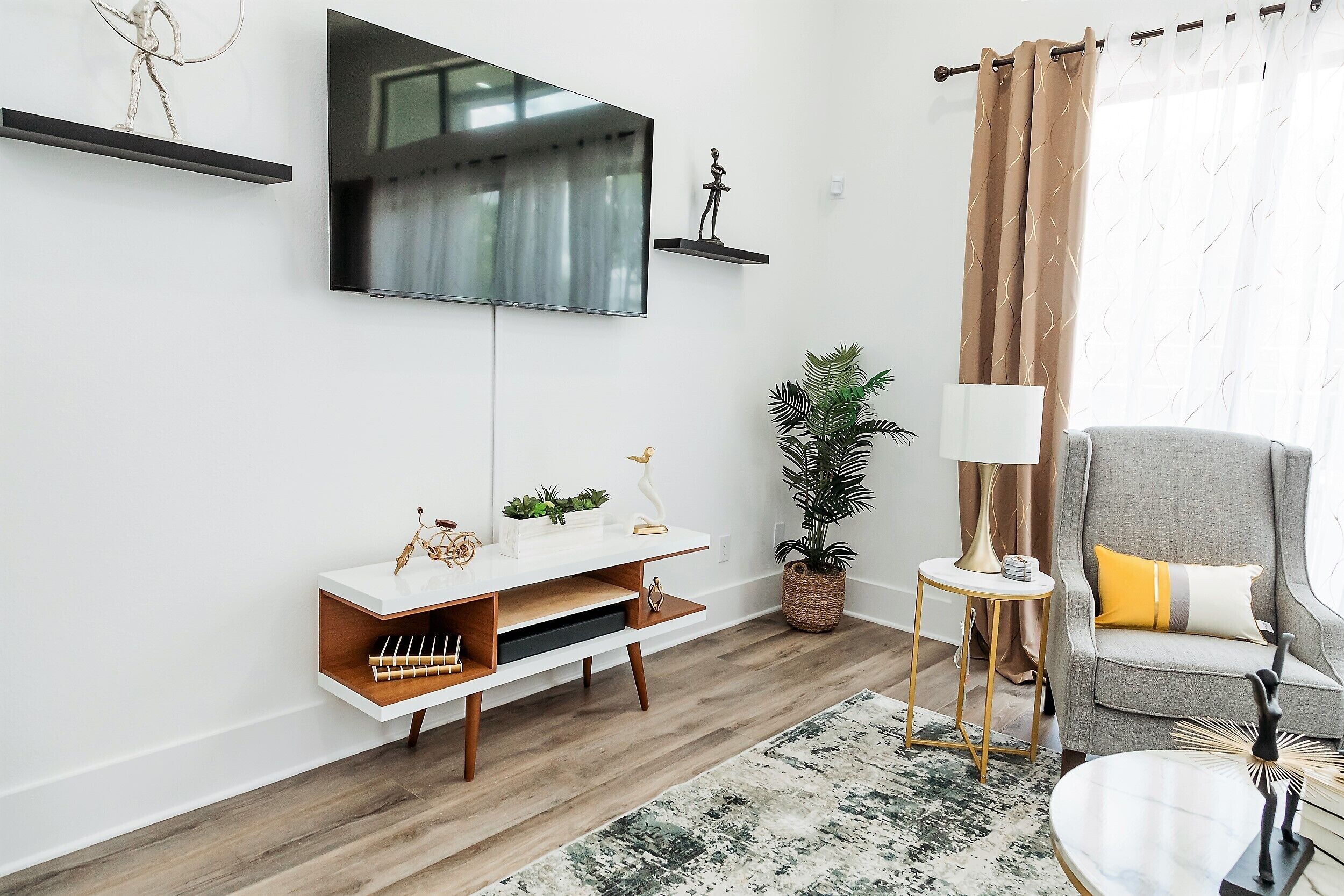 A modern living room with a flat-screen TV mounted on the wall, a white console table with decorative items, a grey armchair with a yellow pillow, and a large window with curtains.