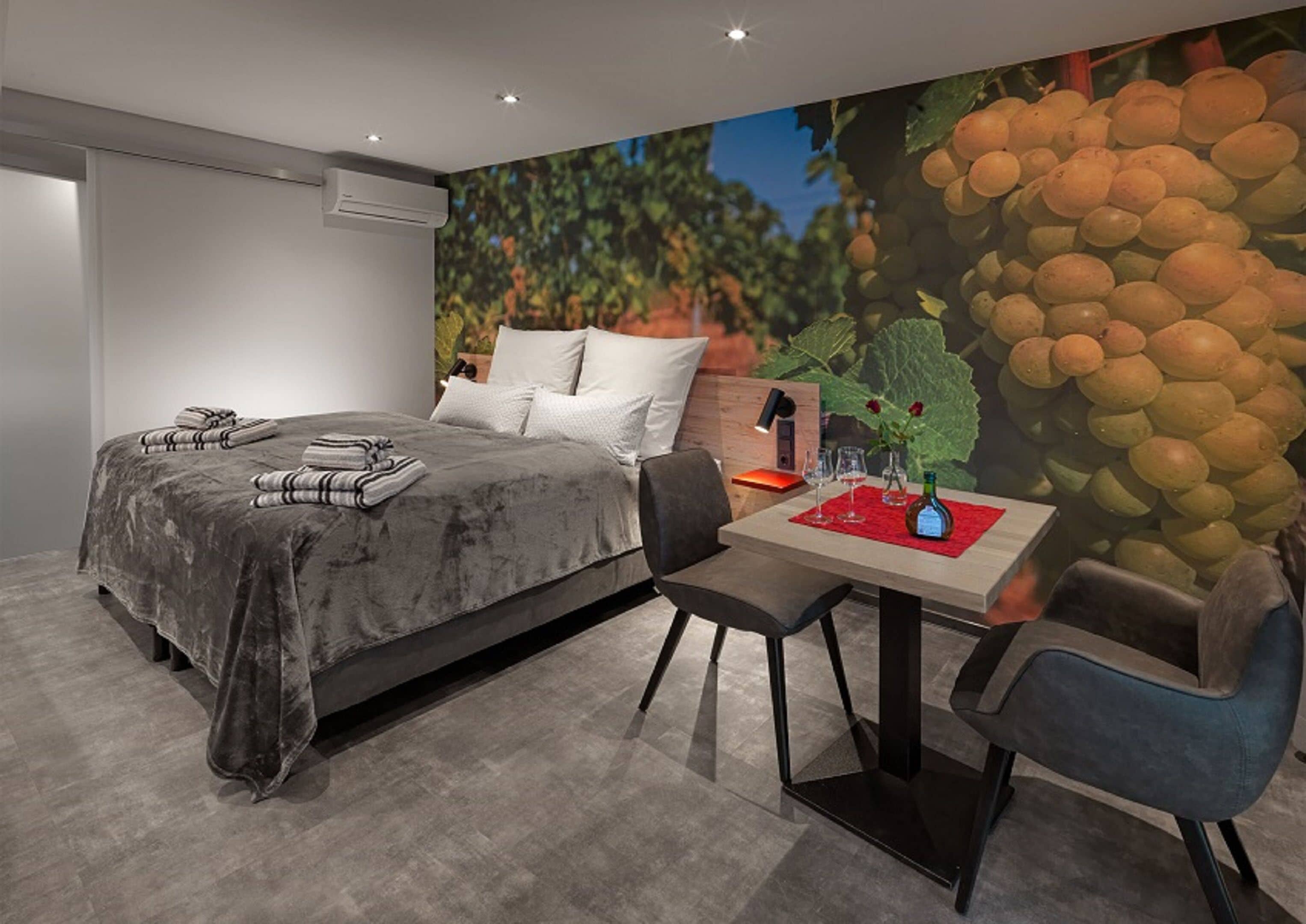 A modern hotel room with a large bed, a dining area with a table and chairs, and a mural of grapes on the wall.