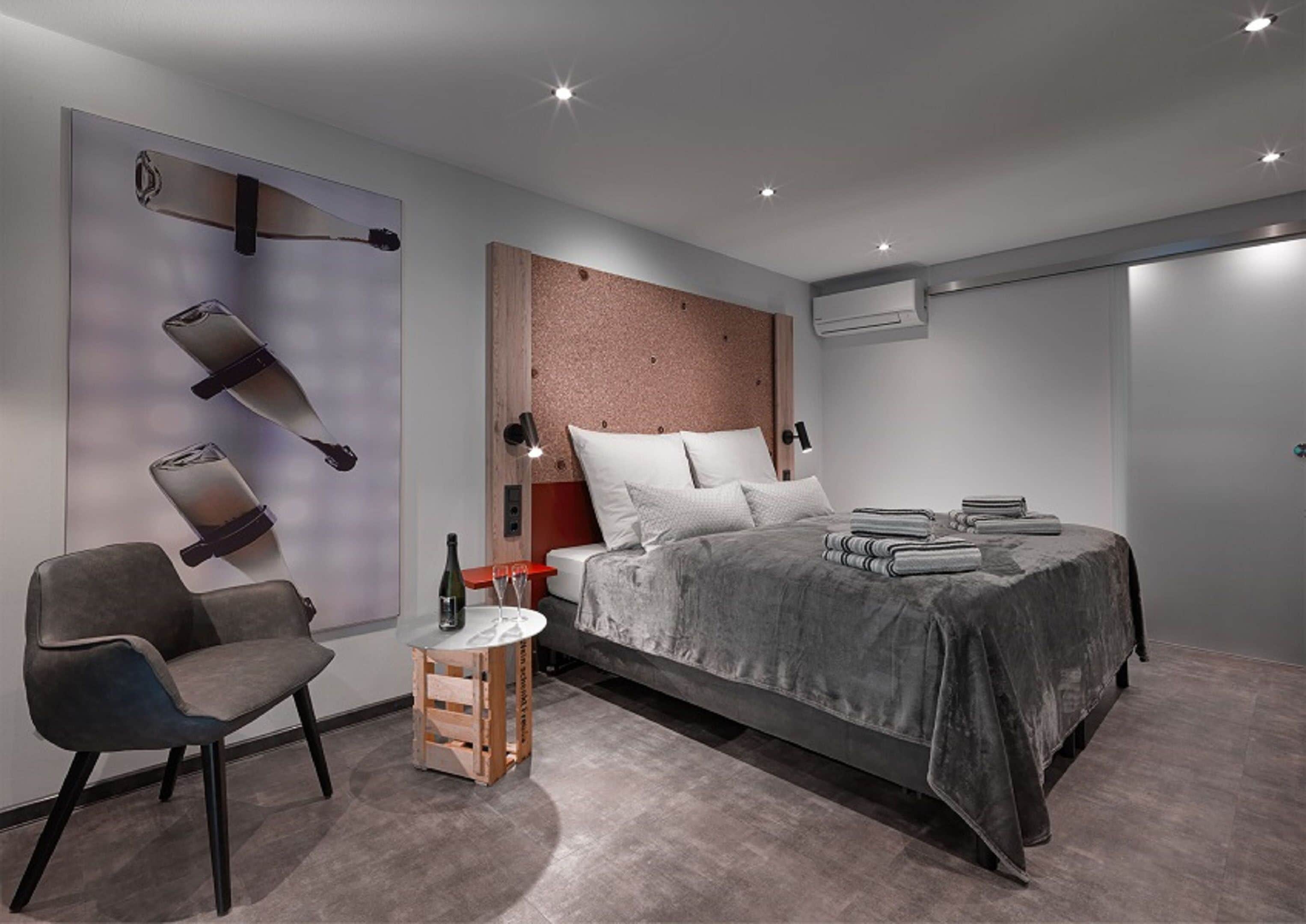 A modern bedroom with a bed, a chair, a small table with a bottle and glasses, and a wall-mounted light fixture.