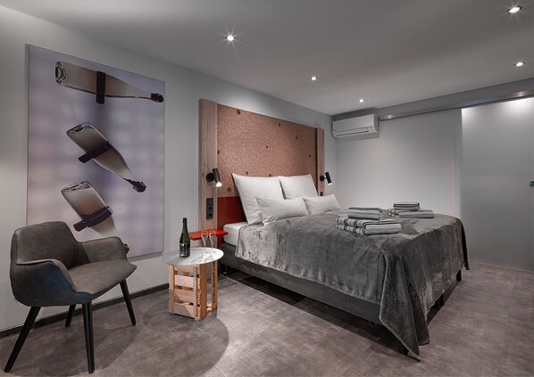 Un dormitorio moderno con una cama, una silla, una mesita con una botella y vasos, y una lámpara de pared.