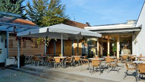 An outdoor café with tables and chairs, covered by umbrellas, and a building entrance with a sign. - WeinMotel Simon 1 a (Alzenau)