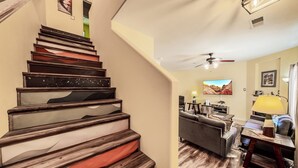 Eine Treppe mit Holzhäuschen und einem roten Teppich, die zu einem Wohnbereich mit einer Couch, einem Couchtisch und einem Fernseher führt.