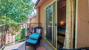 Ein Balkon mit Korbstuhl, kleinem Tisch, einer Schiebeglastür und Blick auf einen Garten sowie benachbarte Gebäude.