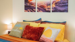 Ein Bett mit bunten Kissen und einem Gemälde, das einen Sonnenuntergang über Felsen zeigt.