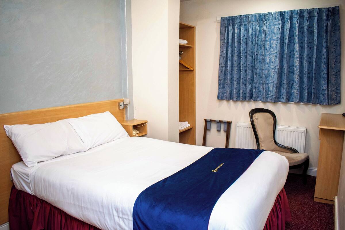 Une chambre d’hôtel avec un lit, une chaise, un bureau, un radiateur et un rideau bleu.