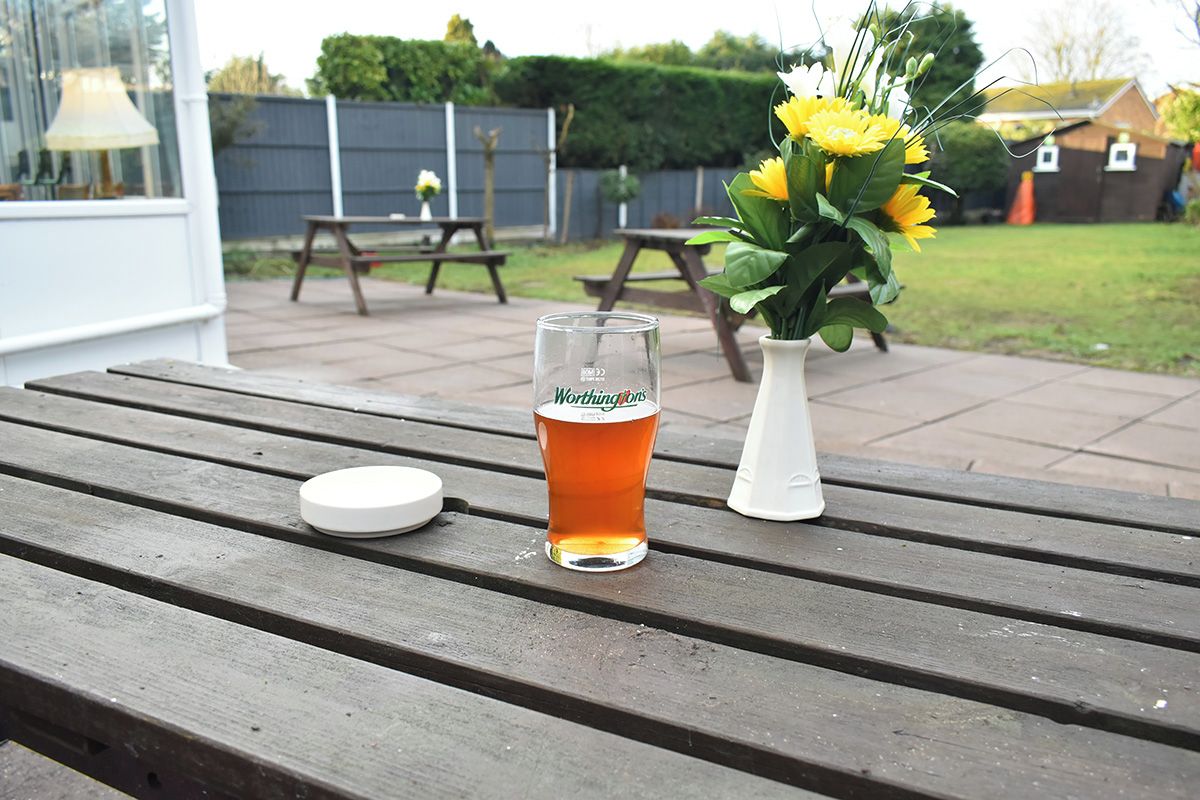 A glass of beer on a wooden table with a white lid beside it, and a white vase with yellow flowers in the background.