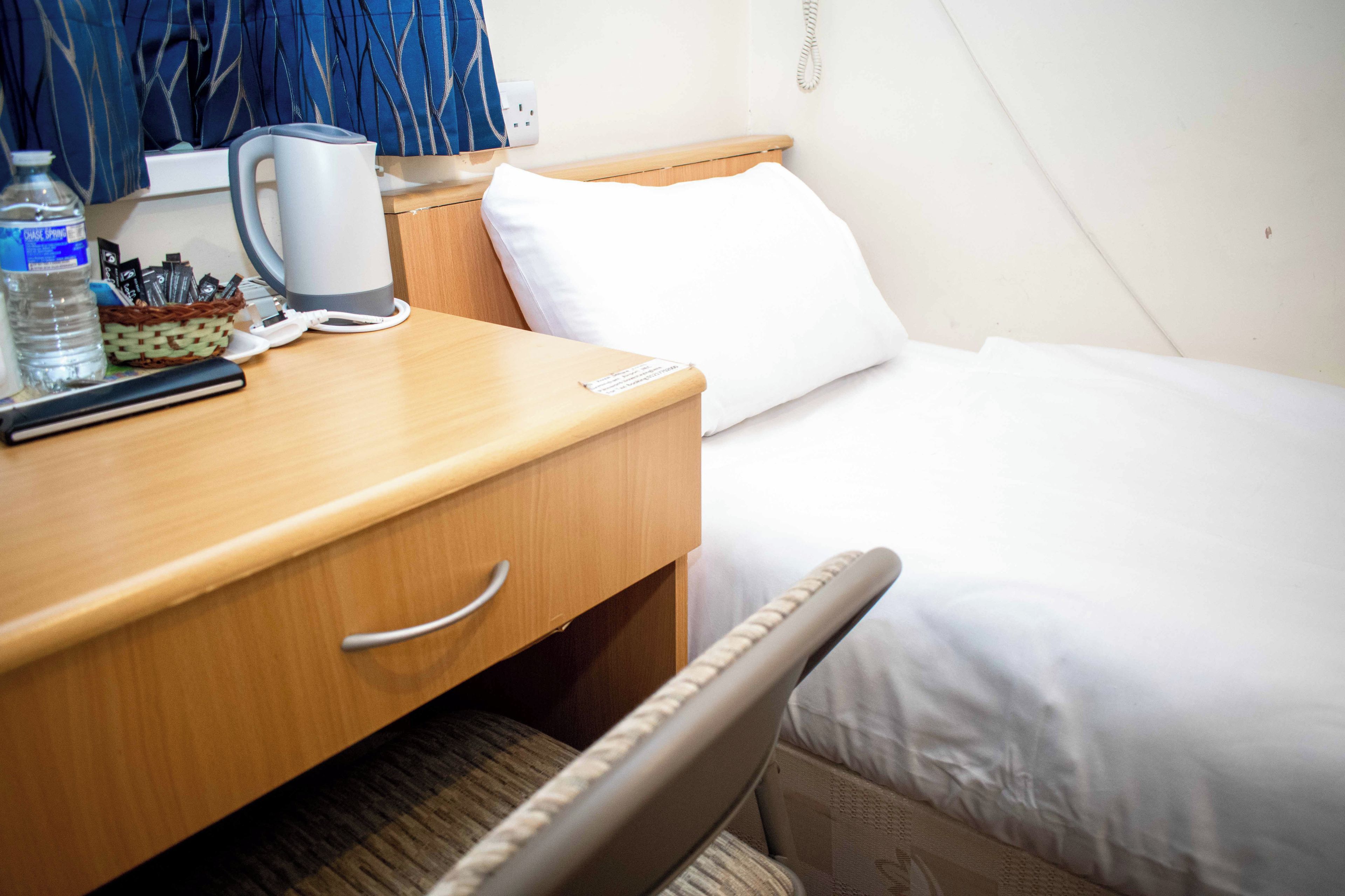 A hotel room with a wooden desk, a kettle, a bottle of water, a basket with pens, a remote control, and a bed with white bedding.