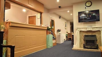 A hotel lobby with a reception desk, a fireplace, a television displaying 'Delhi Crime', and a clock on the wall.