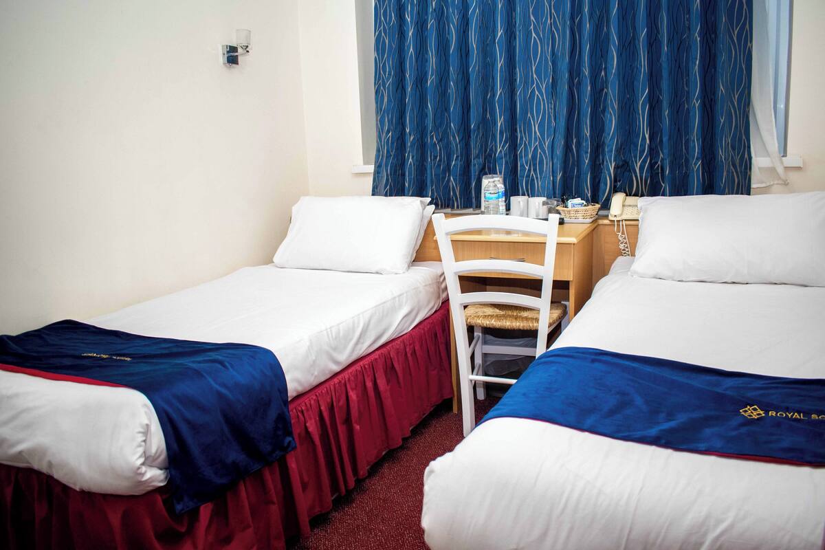 Une chambre d’hôtel avec deux lits simples, un bureau avec une chaise, un téléphone et une fenêtre avec des rideaux bleus.