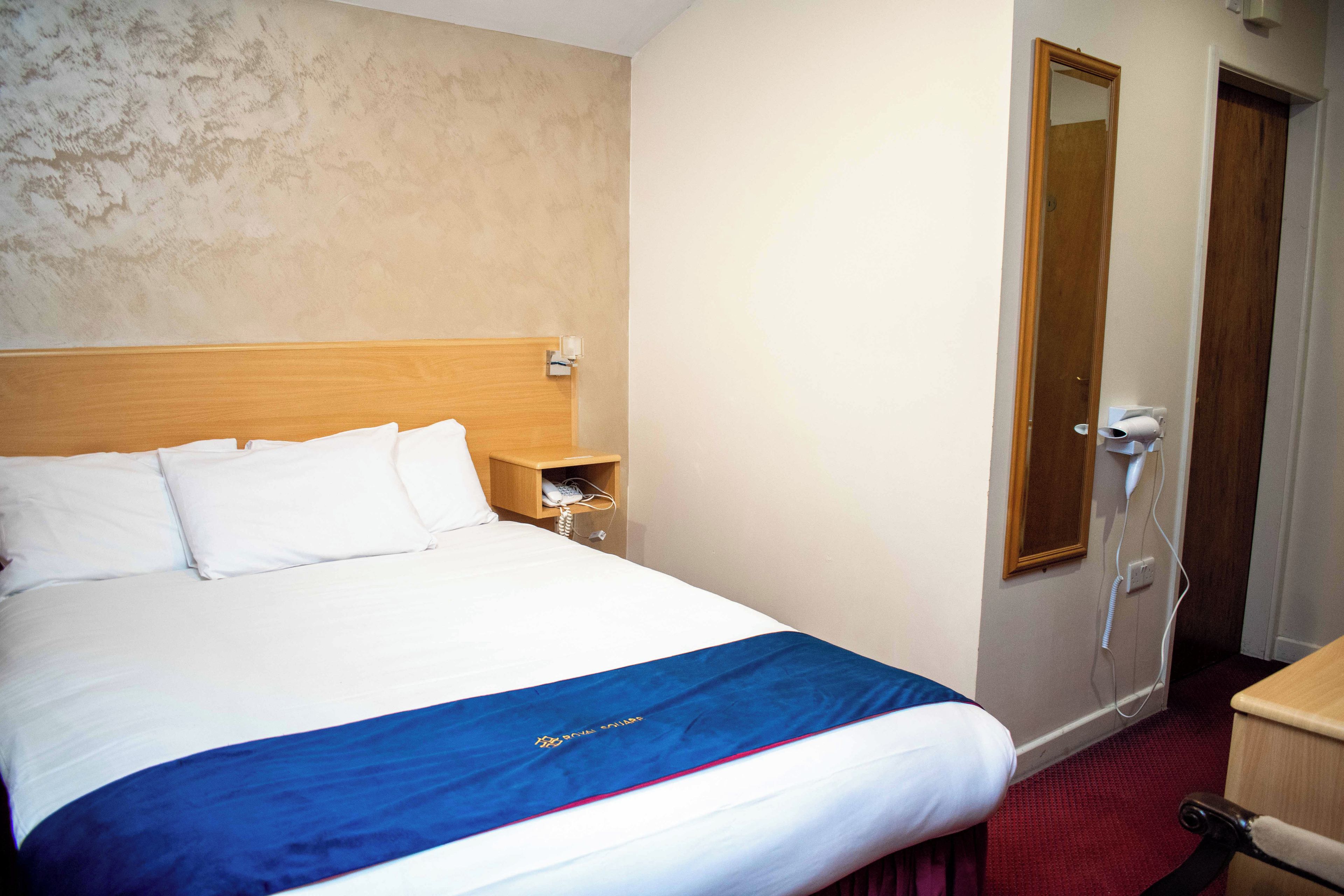 A hotel room with a bed, a wooden headboard, a nightstand, a mirror, and a red carpet.