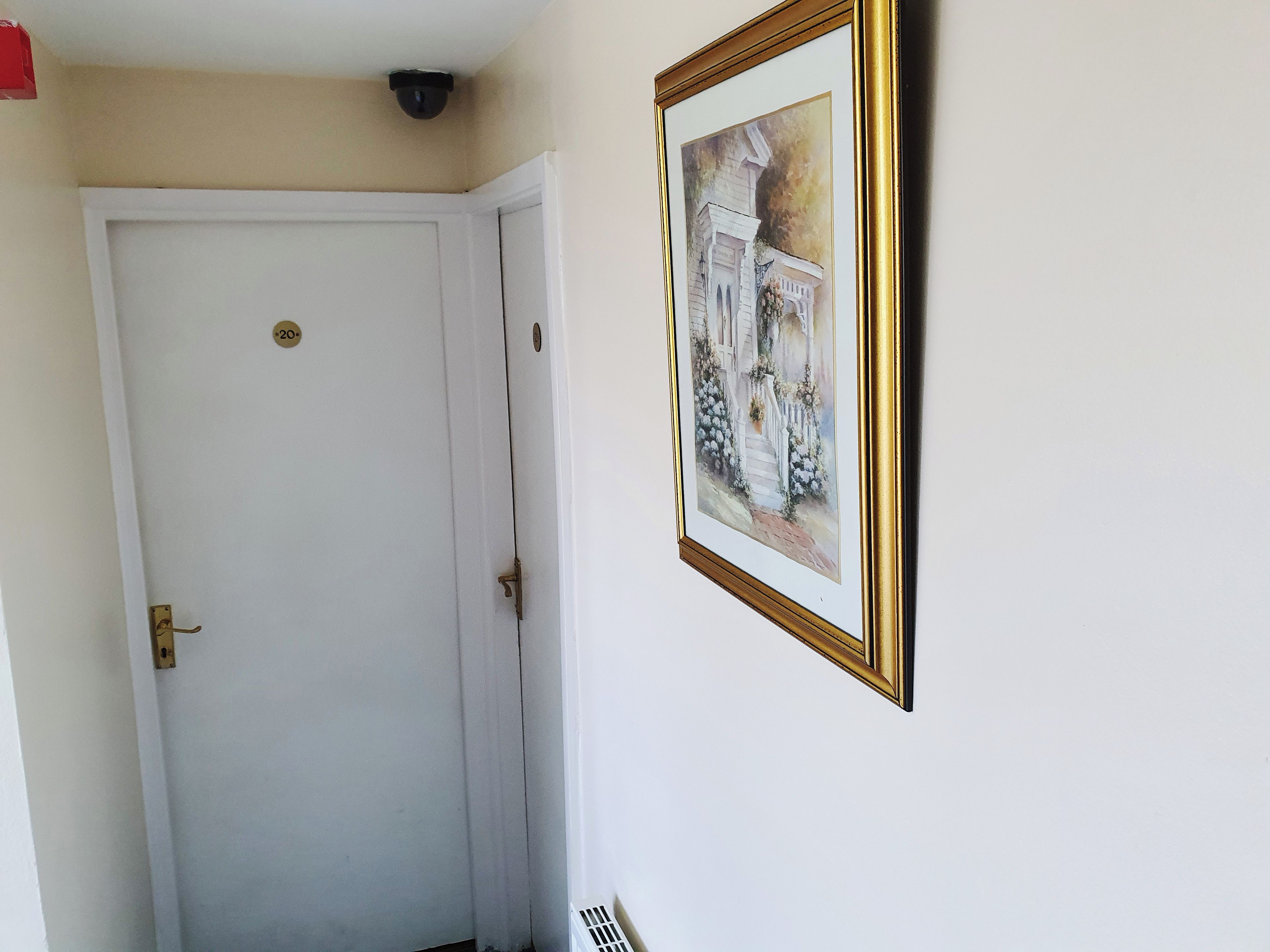 A white door with a brass handle and a number plate, a framed painting of a building with columns, and a vent on the wall.