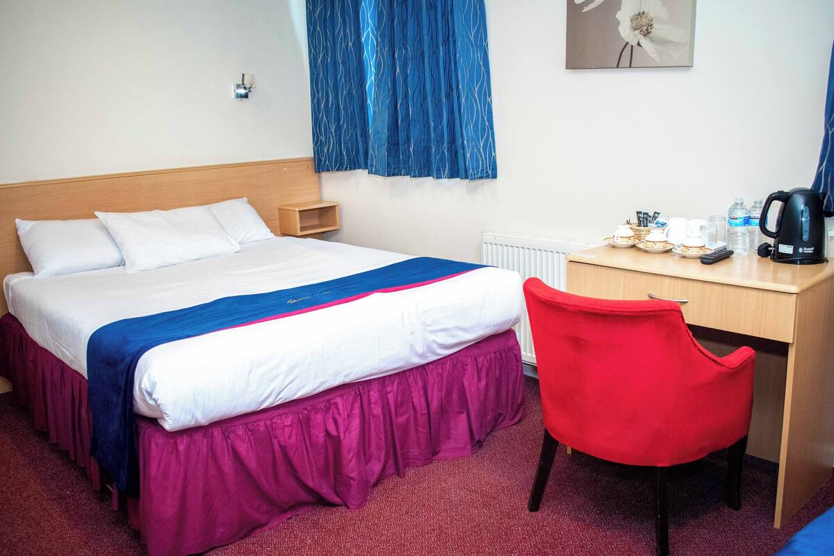 Une chambre d’hôtel comprenant un lit, un bureau, une chaise, un radiateur, une lampe fixée au mur et une fenêtre avec des rideaux.