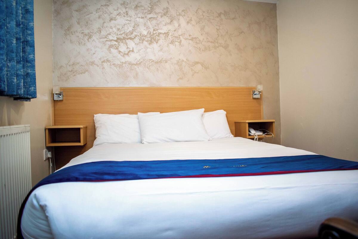 Une chambre d’hôtel avec un lit, une tête de lit en bois, une table de chevet et une lampe fixée au mur.