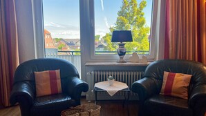Ein Zimmer mit zwei Ledersesseln, einem kleinen Tisch mit Lampe und Blick auf Gebäude und Bäume durch große Fenster.