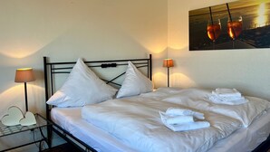Ein ordentlich bezogenes Bett mit weißen Leinen und Kissen, zwei Nachttischlampen und ein Sonnenuntergangsbild an der Wand.