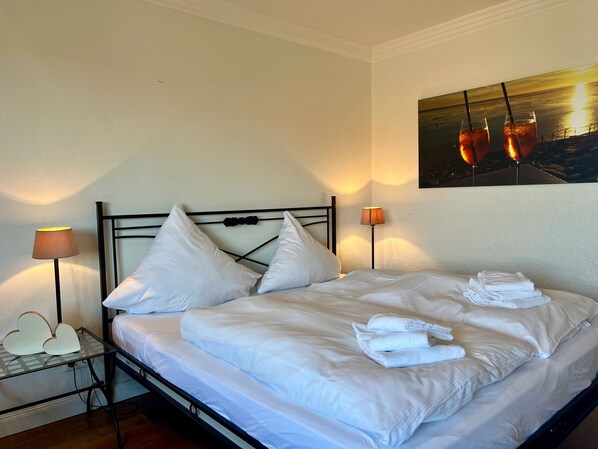 A neatly made bed with white linens and pillows, two bedside lamps, and a painting of a sunset on the wall.