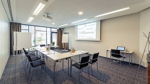 A conference room with a long table, black chairs, a projector screen, and a laptop on a desk. - Altstadthotel Kneitinger (Abensberg)