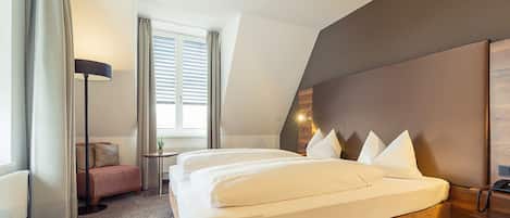 Habitación de hotel moderna con una cama grande, una mesita de noche, una lámpara, un pequeño espacio de descanso y una ventana con persianas.