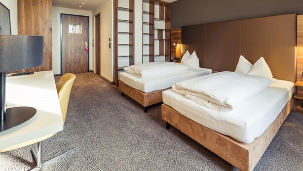 A hotel room with two beds, a desk, and a chair. There is a wooden partition with shelves and a door in the background. - Altstadthotel Kneitinger (Abensberg)